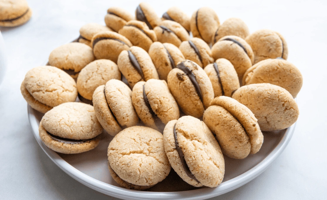 A white plate filled with small, round sandwich cookies with a chocolate filling in the center, set on a white surface.
