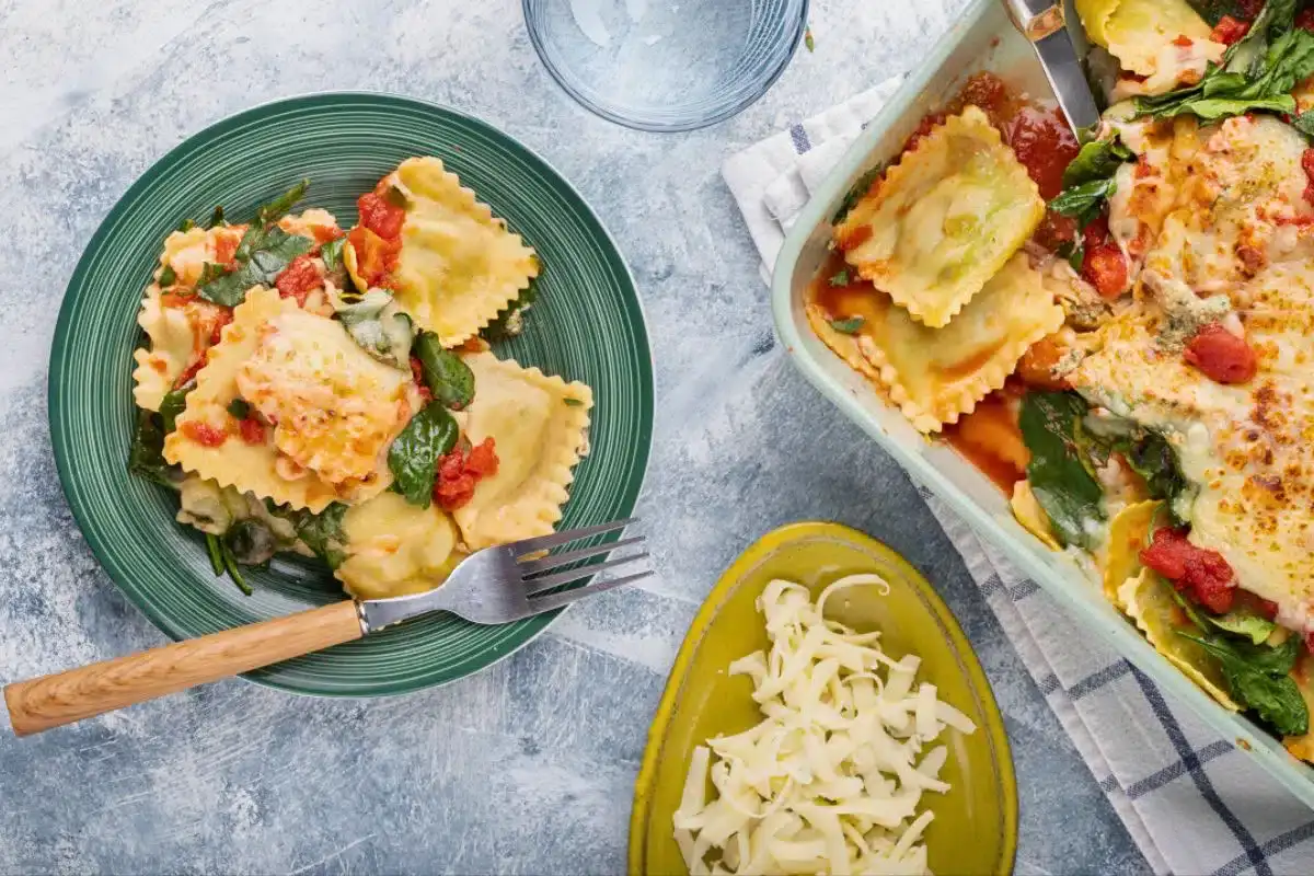 A plate of baked ravioli with spinach and tomato sauce next to a baking dish and a small plate of shredded cheese on a gray surface.