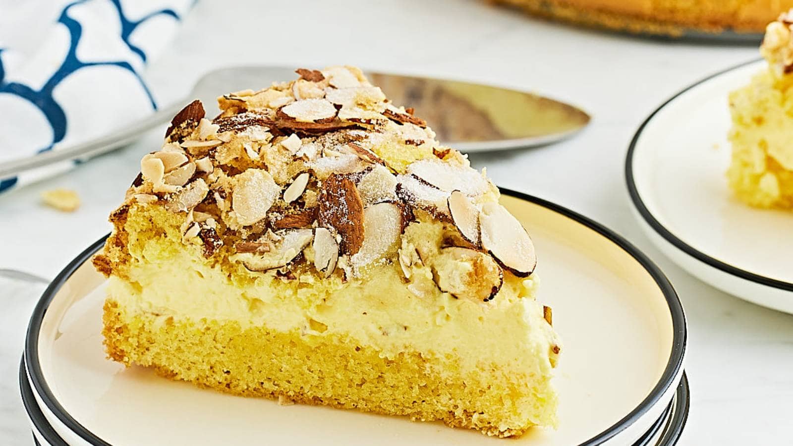 A slice of cake with a yellow sponge base, creamy filling, and topped with sliced almonds sits on a white plate.