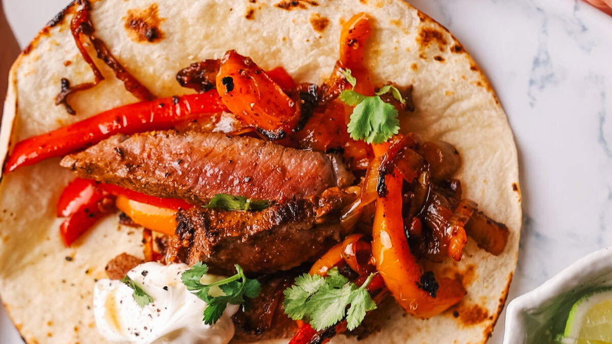 A flour tortilla topped with grilled steak strips, sautรฉed red and orange bell peppers, onions, cilantro, and a dollop of sour cream.