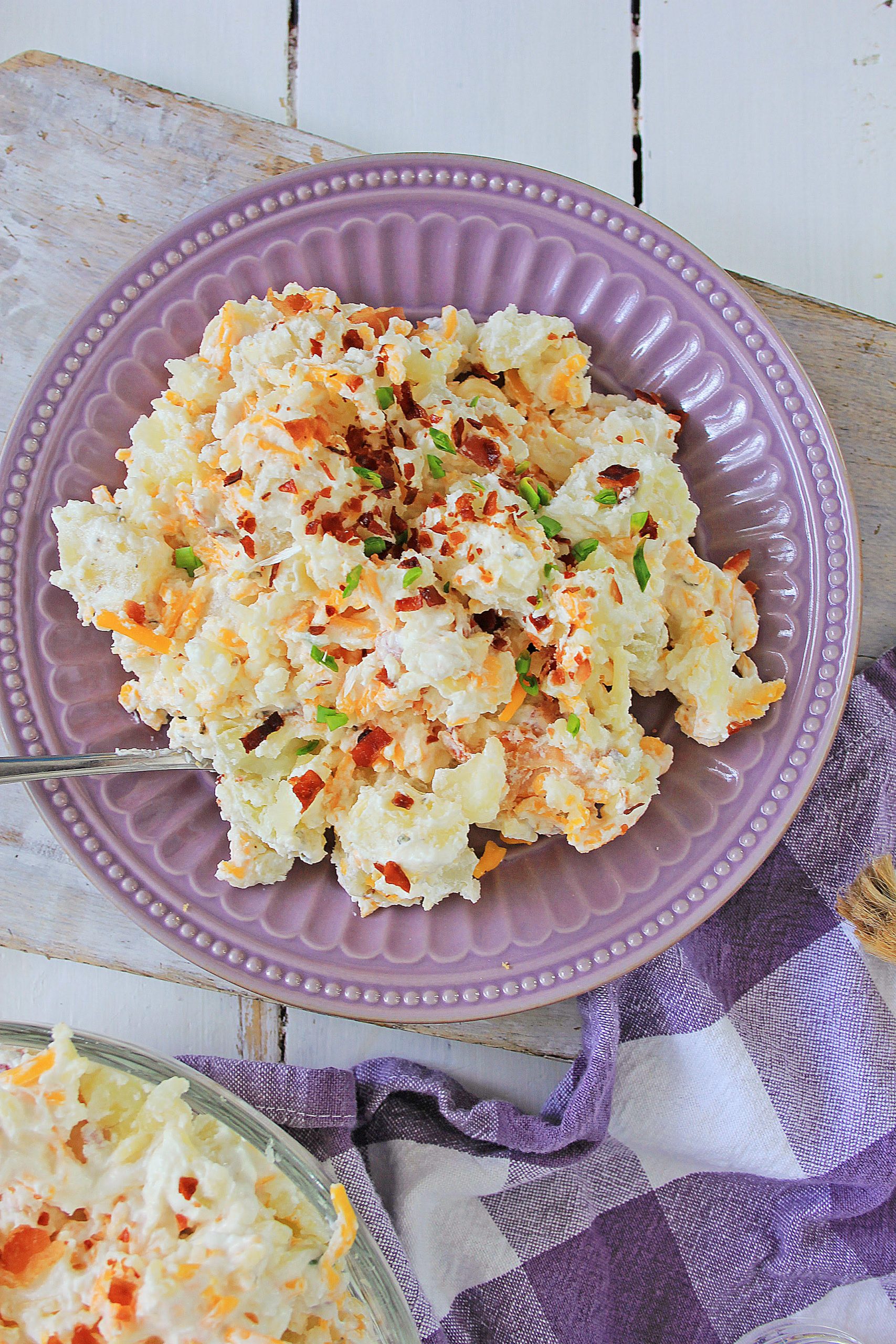 A plate of potato salad topped with chopped green onions and red pepper flakes, served on a purple dish with a checkered napkin underneath.