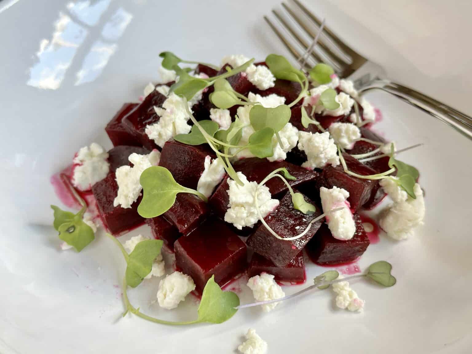 A white plate with diced beet salad topped with crumbled cheese and green microgreens, with a fork placed in the background.