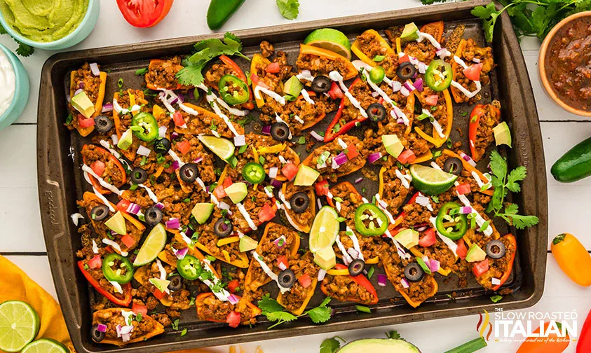 Sheet pan filled with loaded nachos topped with ground meat, sliced peppers, olives, jalapeรฑos, lime wedges, red onion, tomato, cilantro, and drizzled sauce.