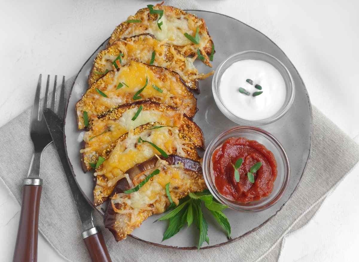 A plate with baked eggplant slices topped with melted cheese, served with two dipping sauces and garnished with fresh herbs. Fork and knife placed beside the plate.