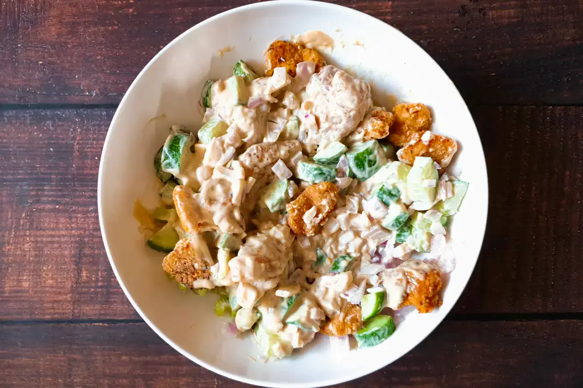 A white bowl containing a salad with greens, breaded chicken pieces, chopped vegetables, and a creamy dressing on a wooden surface.