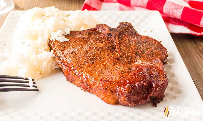 A grilled seasoned pork chop is served next to a portion of white rice on a white plate with a fork.