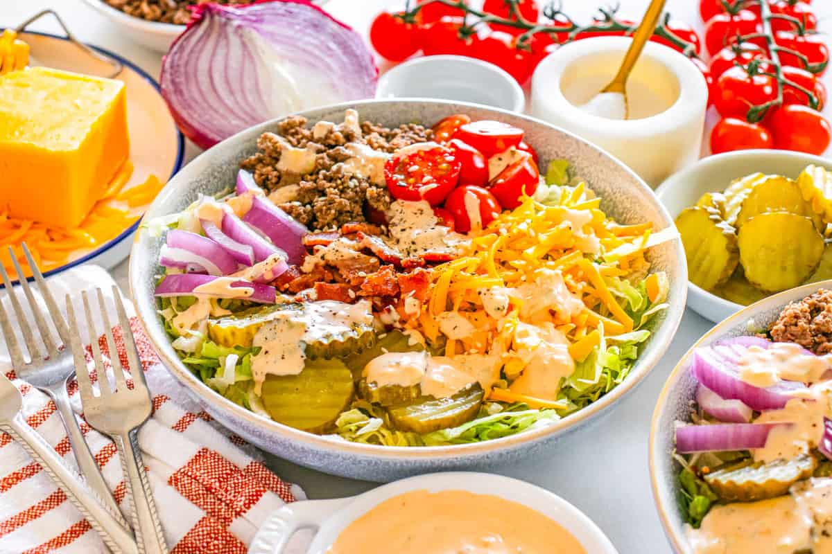 A bowl of salad topped with ground beef, shredded cheese, pickles, red onion, cherry tomatoes, bacon, and creamy dressing, surrounded by salad ingredients and utensils.