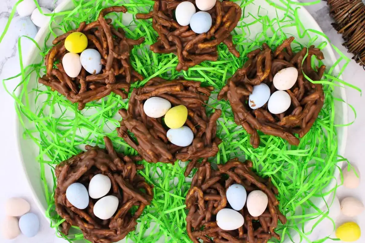 Five chocolate nests made from pretzel sticks are topped with colorful candy eggs and displayed on green shredded paper.