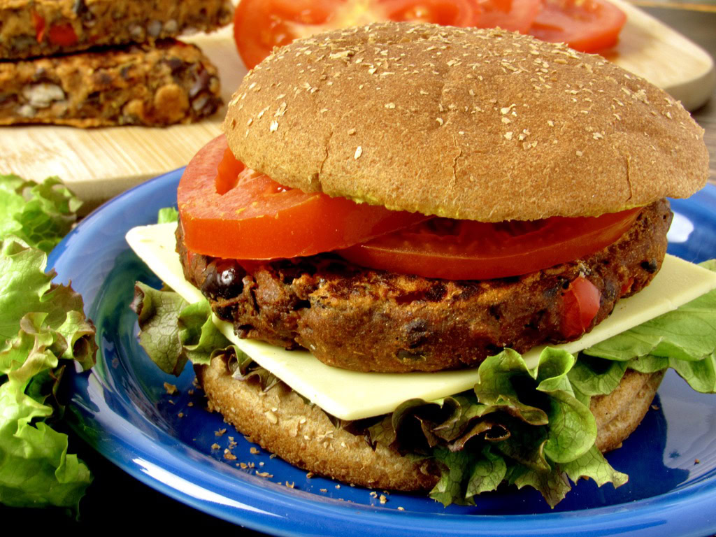 A veggie burger with lettuce, tomato slices, cheese, and a whole wheat bun is served on a blue plate, with extra tomato slices in the background.