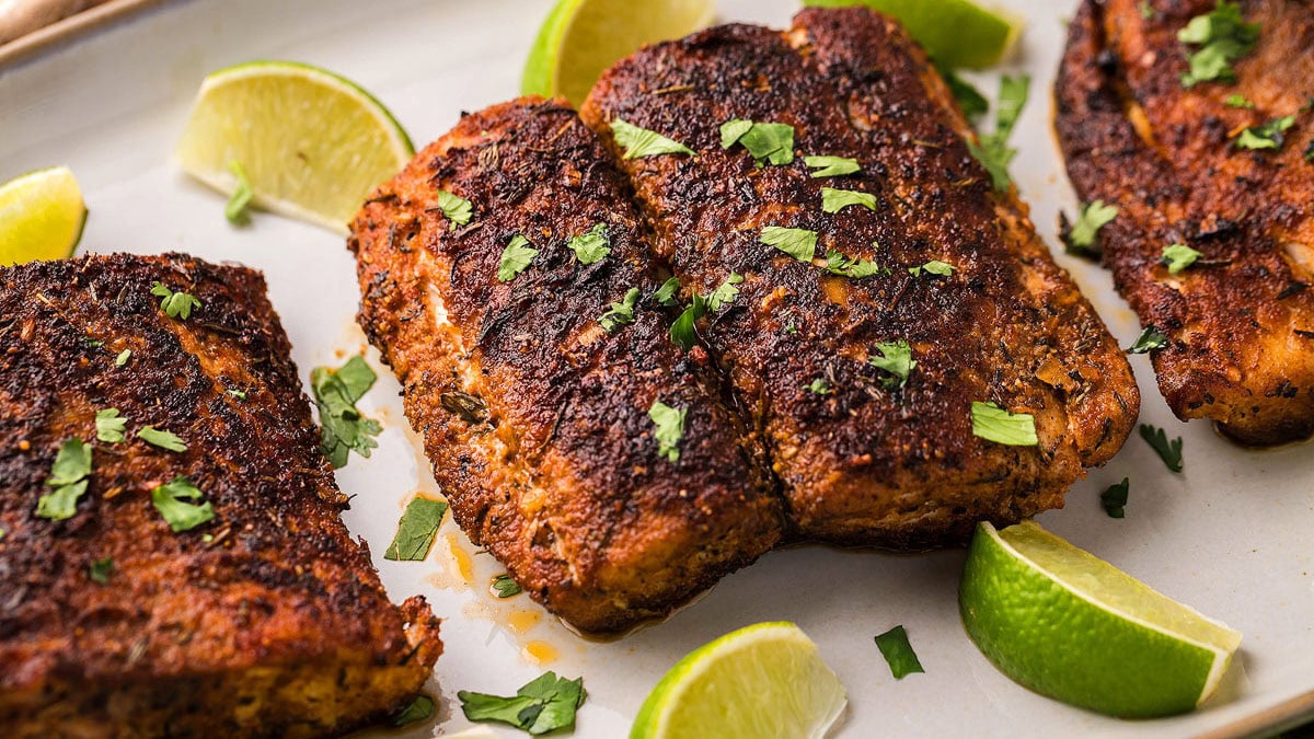 Two pieces of blackened fish garnished with chopped herbs, served on a plate with lime wedges.