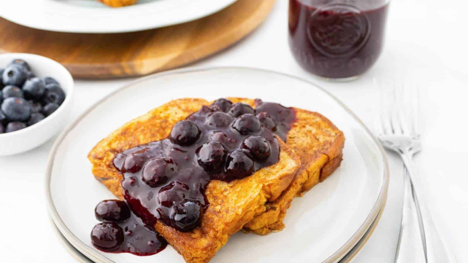 Two slices of French toast topped with blueberry compote on a white plate, with a bowl of blueberries, a jar of compote, and forks nearby.