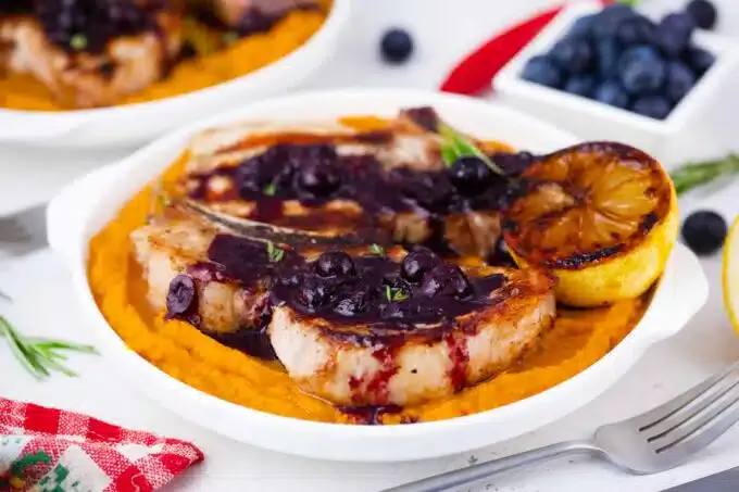 Grilled pork chops with blueberry sauce and a grilled lemon slice served over a bed of orange mashed sweet potatoes.