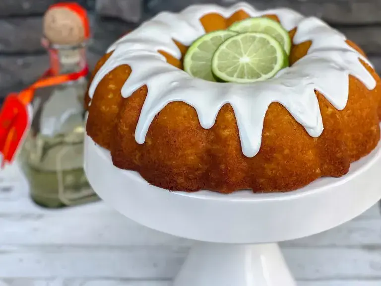 A Bundt cake with white icing and lime slices on top sits on a white cake stand; a bottle of tequila is in the background.