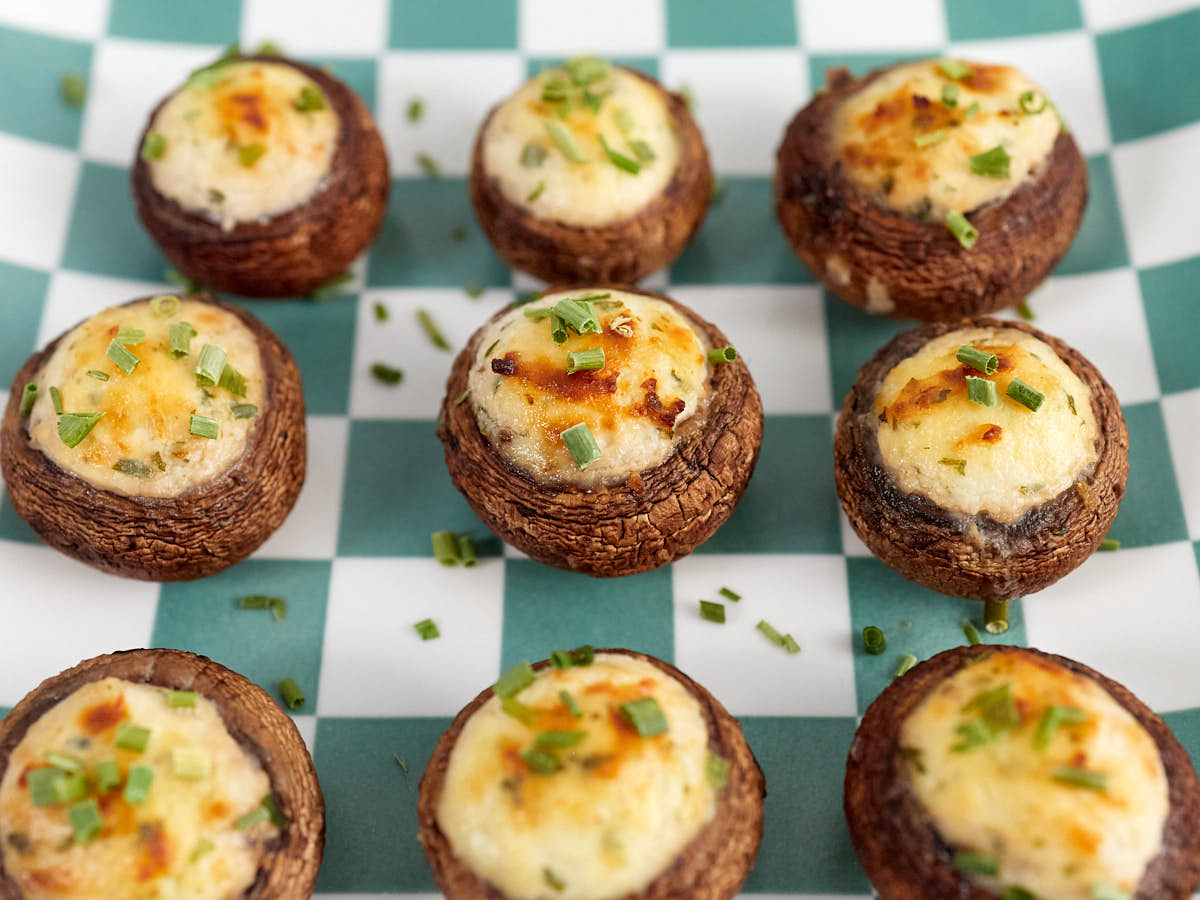 Nine stuffed mushrooms topped with chopped chives are arranged on a green and white checkered surface.