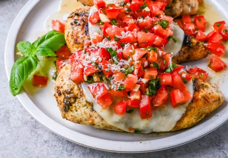 Grilled chicken breasts topped with melted cheese, diced tomatoes, herbs, and grated parmesan, served on a white plate with a basil garnish.