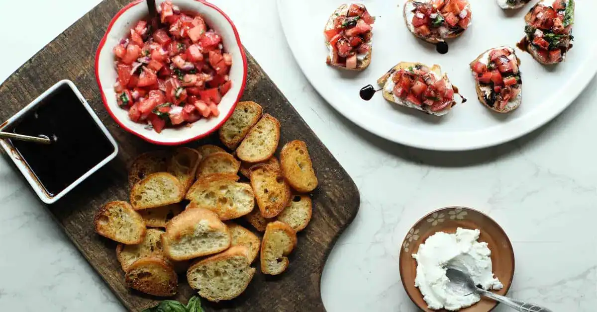 Sliced toasted bread, a bowl of tomato bruschetta mix, balsamic vinegar, and a small dish of cheese are arranged on a wooden and marble surface.