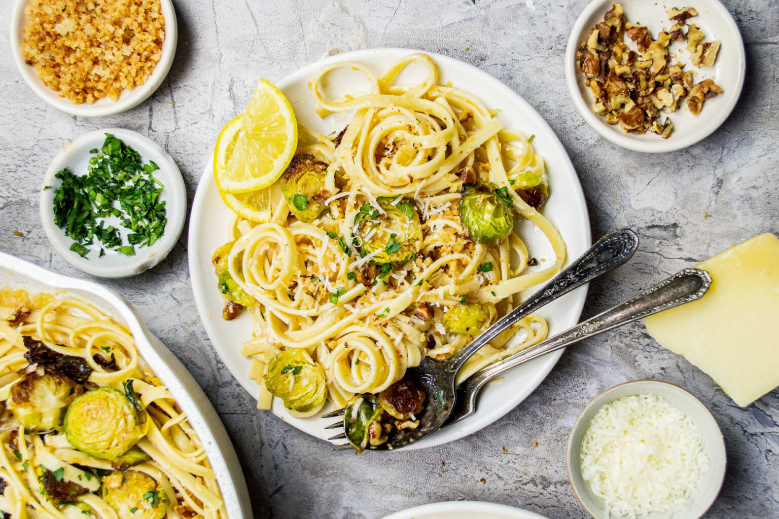 Plate of linguine pasta with Brussels sprouts, lemon wedges, and herbs, surrounded by bowls of chopped nuts, parsley, grated cheese, and breadcrumbs on a gray surface.