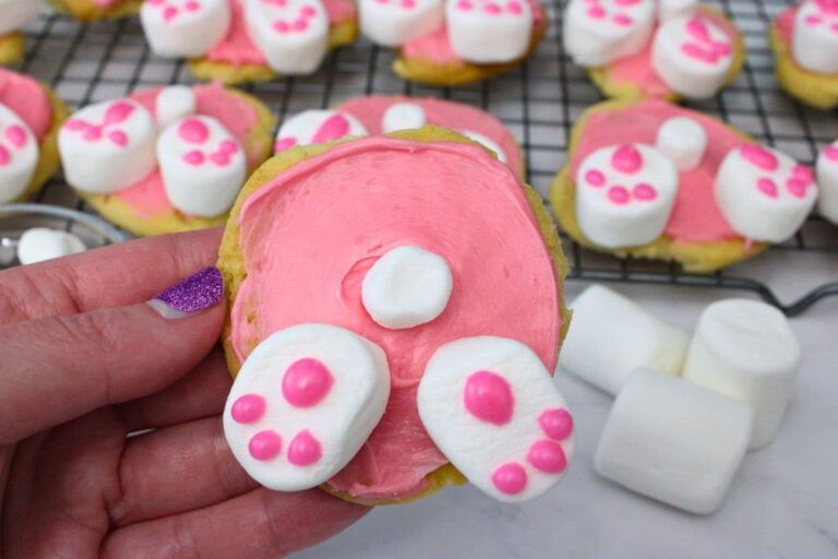A hand holds a cookie decorated with pink icing, marshmallows, and pink dots to resemble a bunny’s hindquarters; more similarly decorated cookies are on a cooling rack in the background.