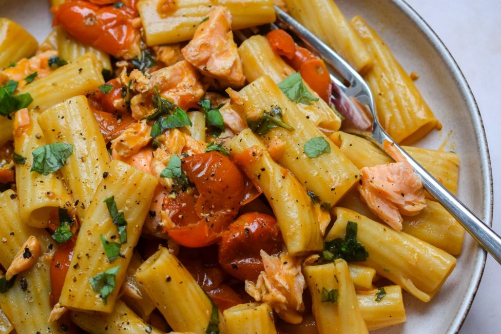 A plate of rigatoni pasta with cherry tomatoes, pieces of salmon, fresh herbs, and black pepper, served with a spoon.