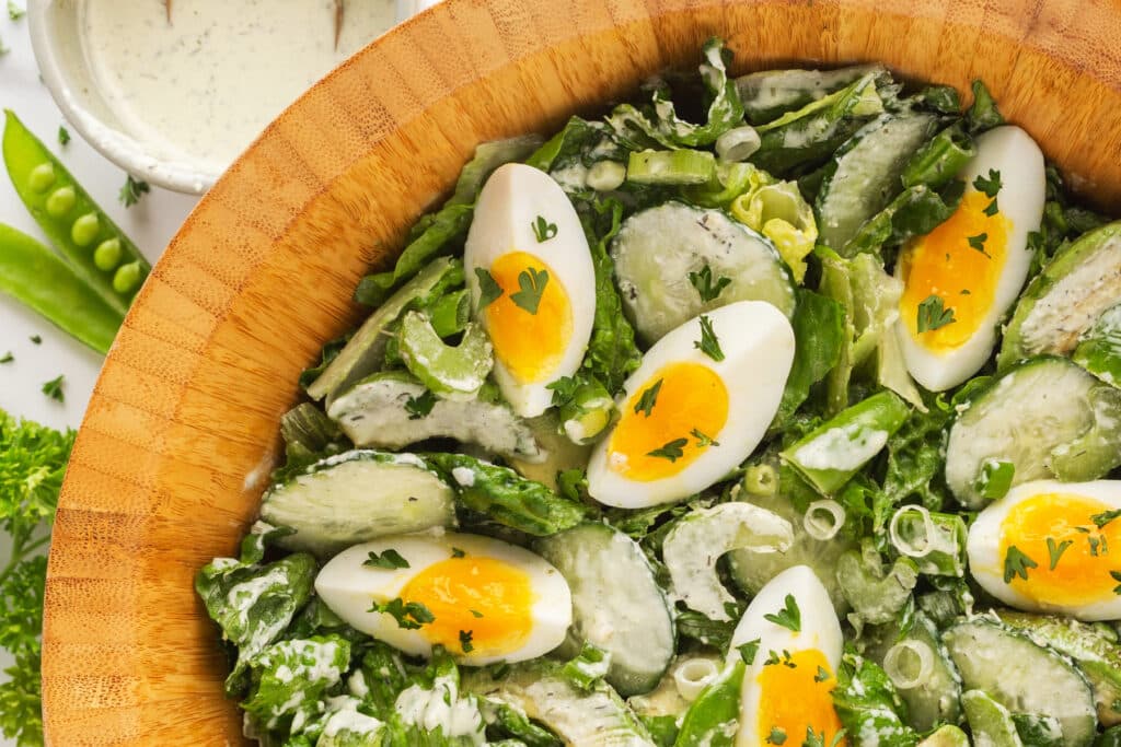 A wooden bowl filled with salad containing lettuce, cucumber slices, snap peas, and quartered hard-boiled eggs, topped with creamy dressing and chopped herbs.