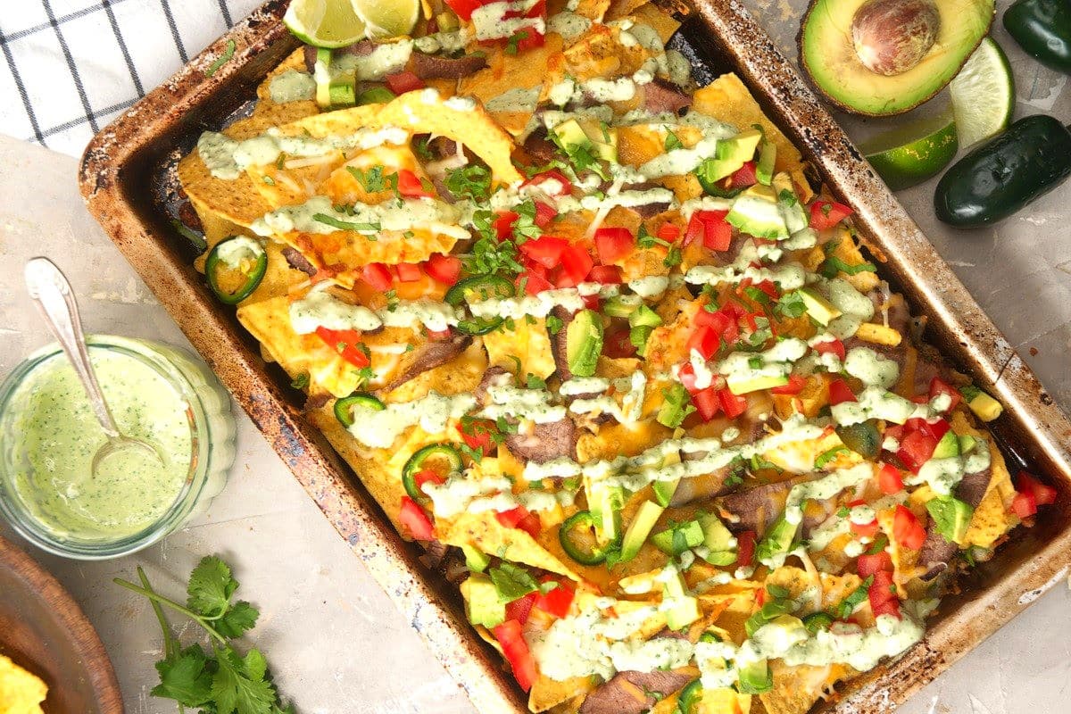 A baking tray filled with nachos topped with diced tomatoes, avocado, jalapeños, cilantro, and a drizzle of creamy green sauce. Lime wedges and an avocado are nearby.