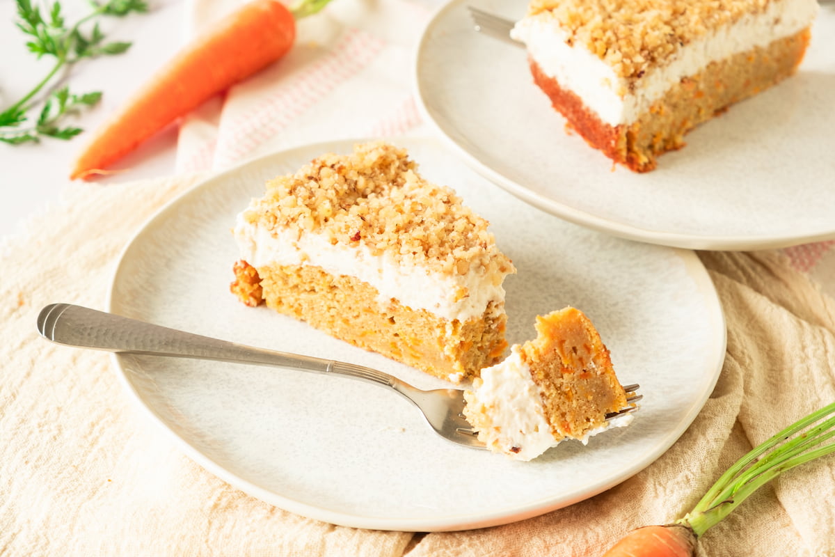 A slice of carrot cake with cream cheese frosting and crumb topping on a plate, with a fork holding a bite. Another slice and carrot are visible in the background.