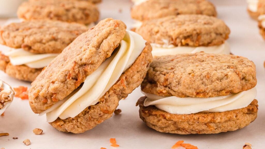 Oatmeal sandwich cookies filled with white cream frosting are arranged on a light surface, with small pieces of carrot visible in the cookies.