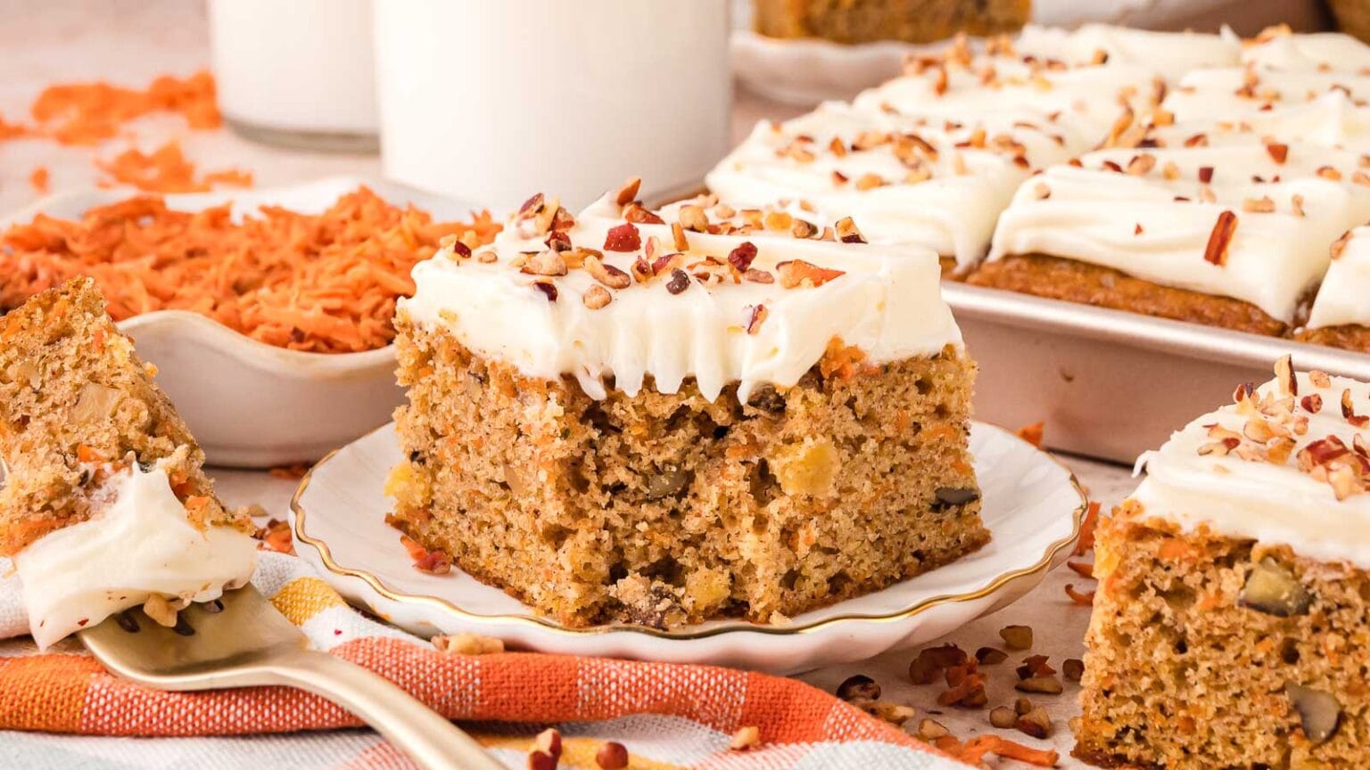 A slice of carrot cake with cream cheese frosting and chopped nuts on a plate, surrounded by more cake pieces and a bowl of grated carrots.