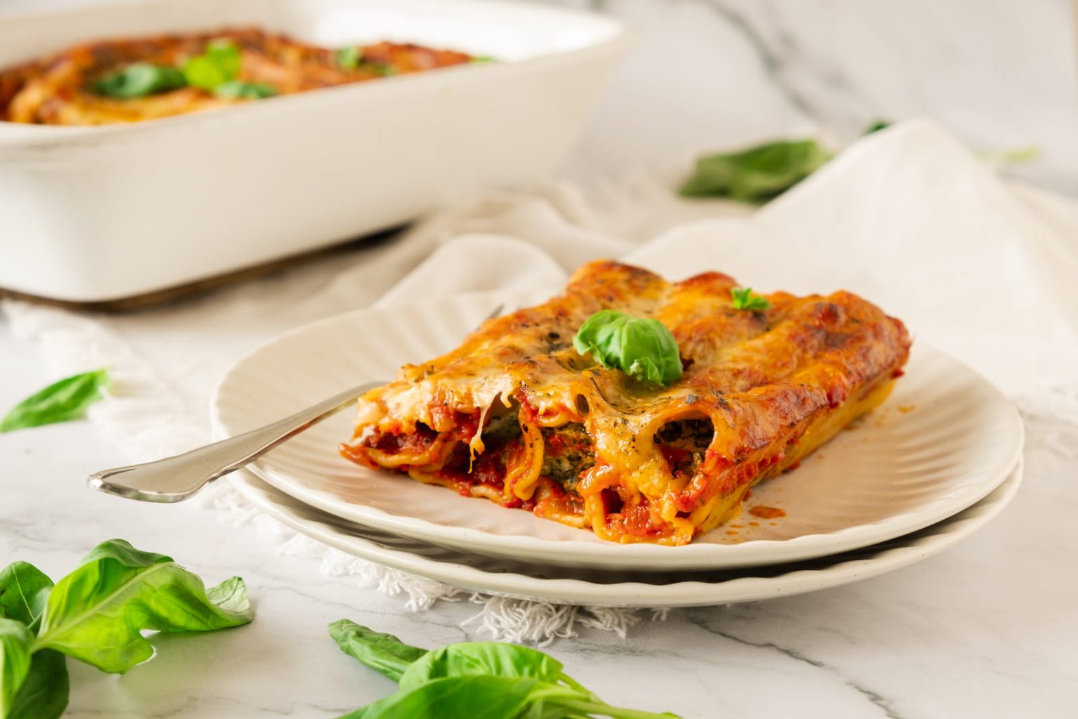A plate with two pieces of baked lasagna garnished with basil, set on a white table with a fork and a baking dish of lasagna in the background.