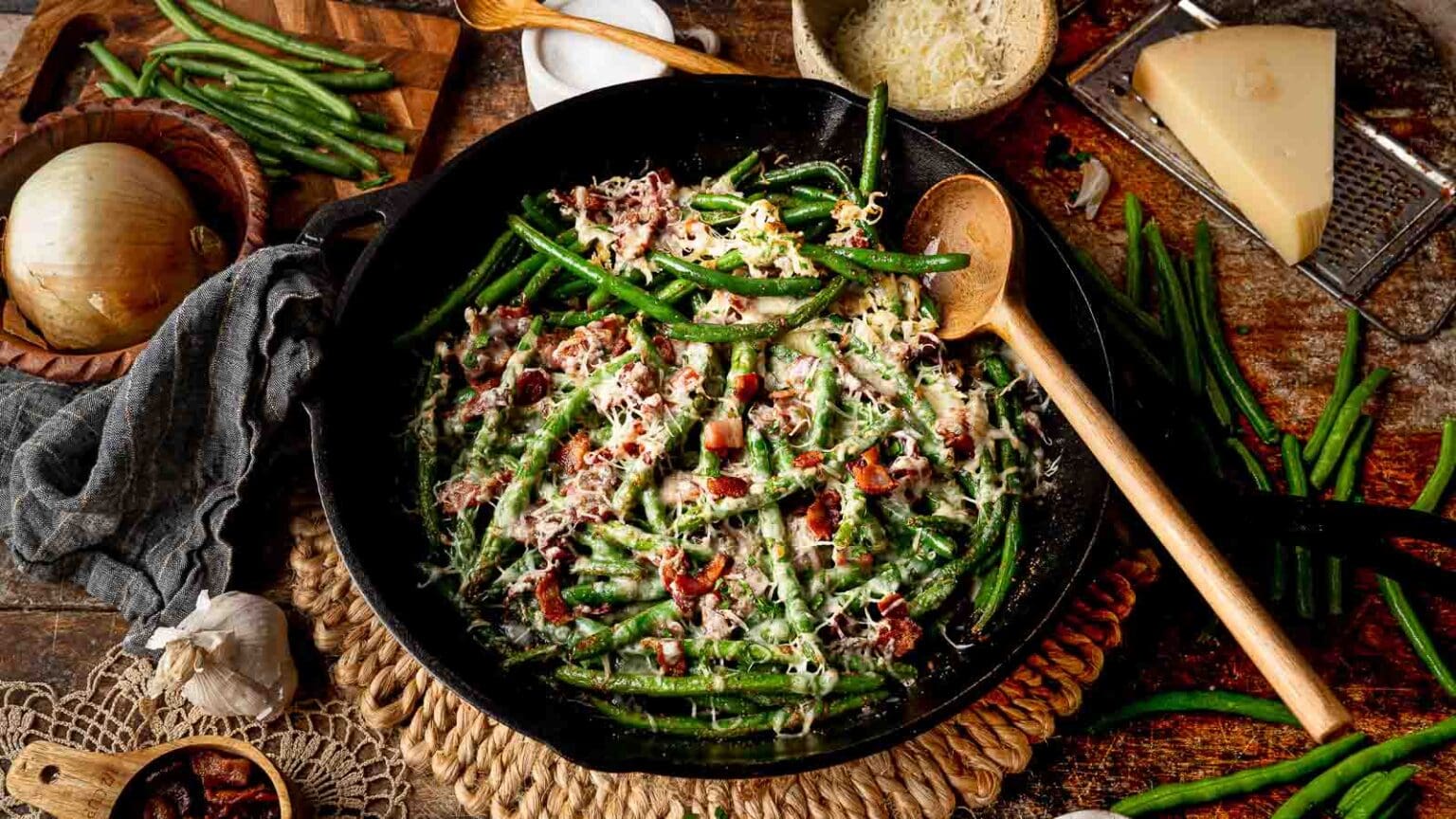 A cast iron skillet filled with green beans, bacon bits, and grated cheese sits on a table surrounded by fresh ingredients, including onion, garlic, green beans, and a wedge of cheese.