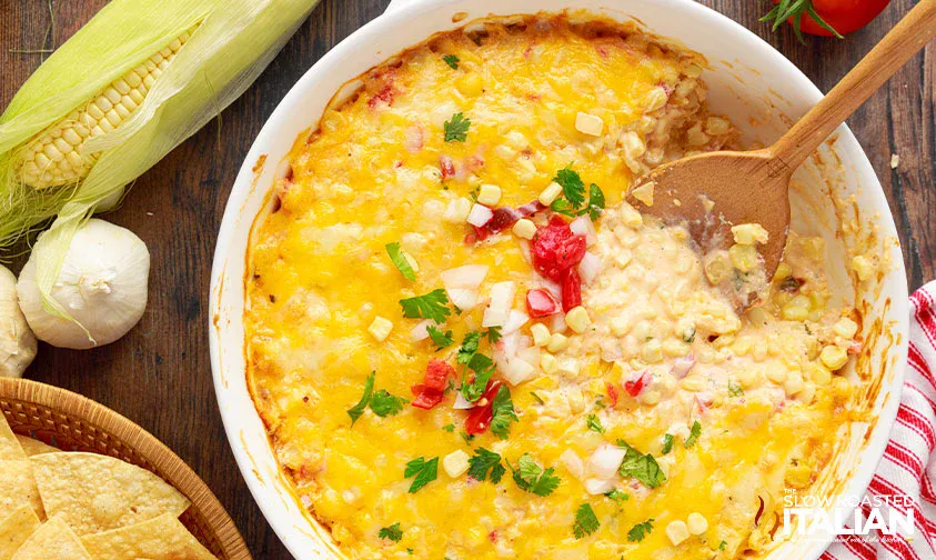 A baked cheesy corn dip in a round white dish, topped with chopped tomatoes, onions, and parsley. A wooden spoon is scooping some dip. Corn, garlic, and chips are nearby.
