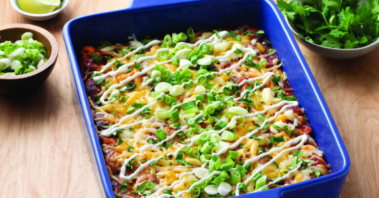 A baked casserole topped with melted cheese, chopped green onions, cilantro, and drizzled with a white sauce in a blue dish. Bowls of lime, green onions, and cilantro are beside it.