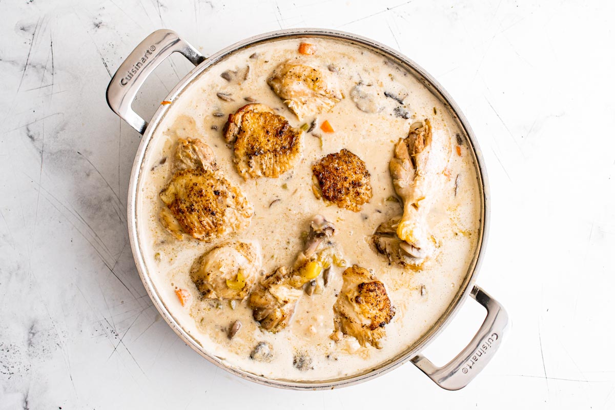 A braised chicken dish with golden brown pieces of chicken in a creamy mushroom sauce, cooked in a large silver pan on a white surface.
