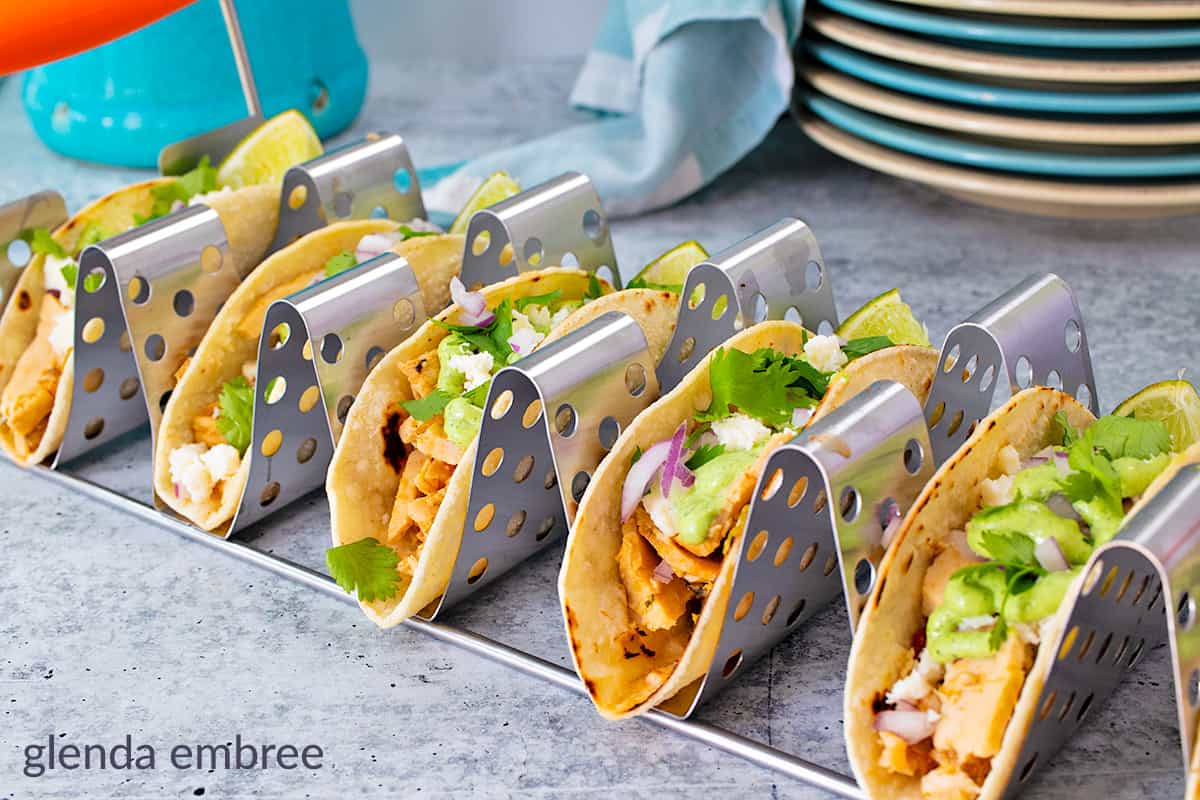 A row of tacos in metal holders, filled with chicken, lettuce, avocado, cheese, cilantro, and onion, set on a gray countertop with plates and a towel in the background.