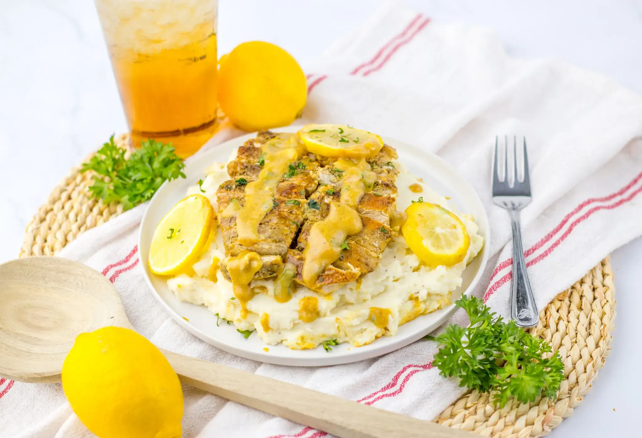 Grilled chicken breast with creamy lemon sauce on mashed potatoes, garnished with lemon slices and parsley, served with iced tea.