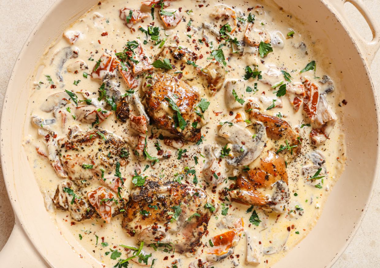 Chicken thighs simmered in a creamy sauce with mushrooms, sun-dried tomatoes, herbs, and black pepper, served in a skillet.