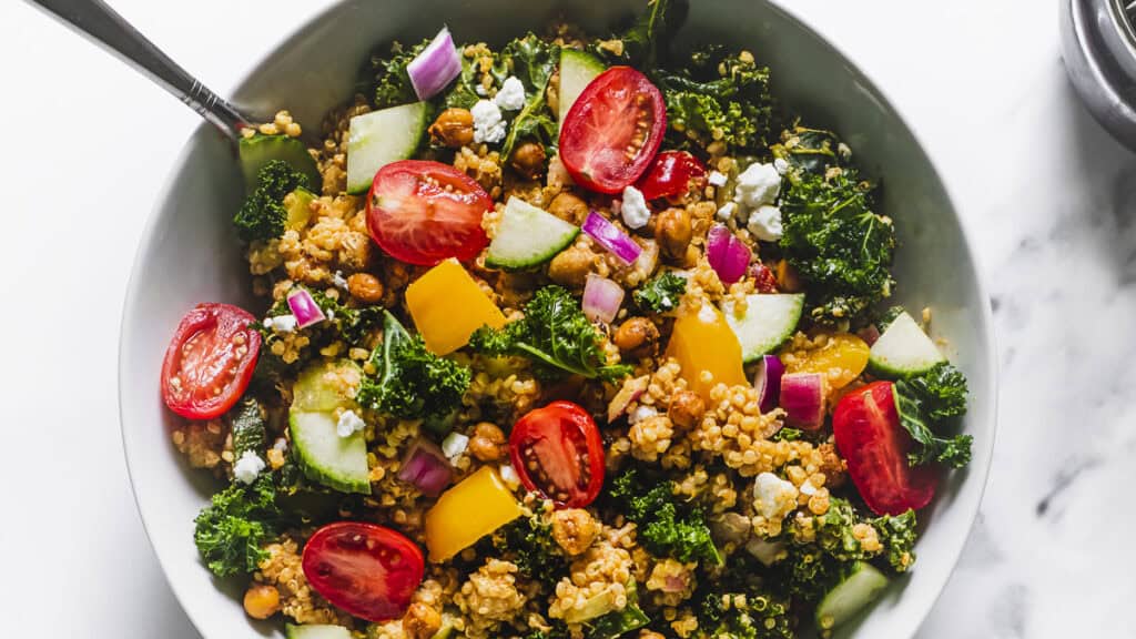 A bowl filled with kale, cherry tomatoes, cucumber, yellow bell pepper, red onion, chickpeas, quinoa, and crumbled cheese, with a fork on the side.