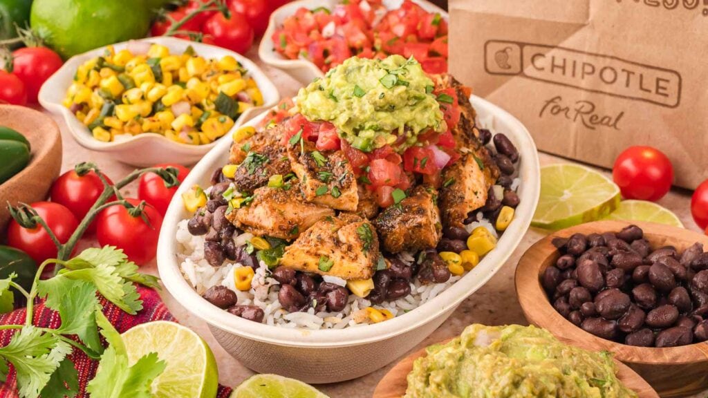 A Chipotle burrito bowl with grilled chicken, black beans, rice, corn, salsa, and guacamole, surrounded by bowls of beans, corn salsa, guacamole, tomatoes, limes, and cilantro.