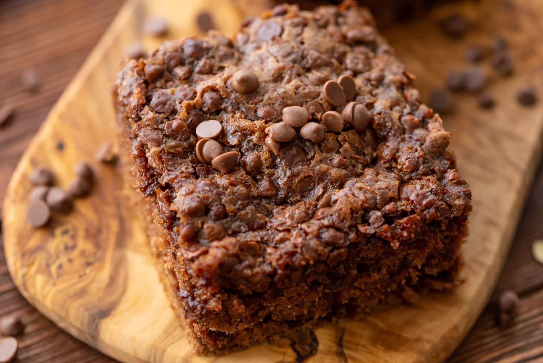 A close-up of a chocolate chip oatmeal bar on a wooden board, topped with chocolate chips and surrounded by a few scattered chips.