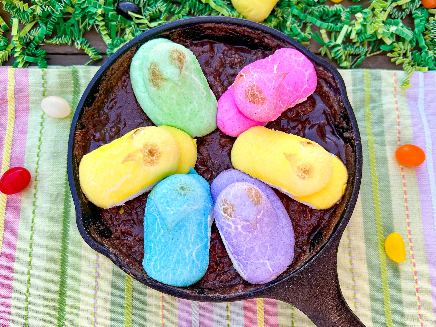 A cast iron skillet holds a chocolate dessert topped with five colorful marshmallow chick candies, surrounded by festive decorations and jelly beans.
