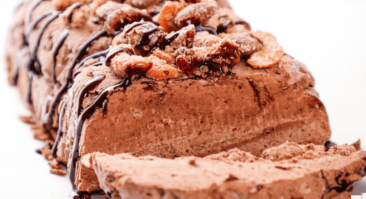Chocolate ice cream loaf topped with nuts and drizzled with chocolate sauce, with a sliced piece in the foreground.