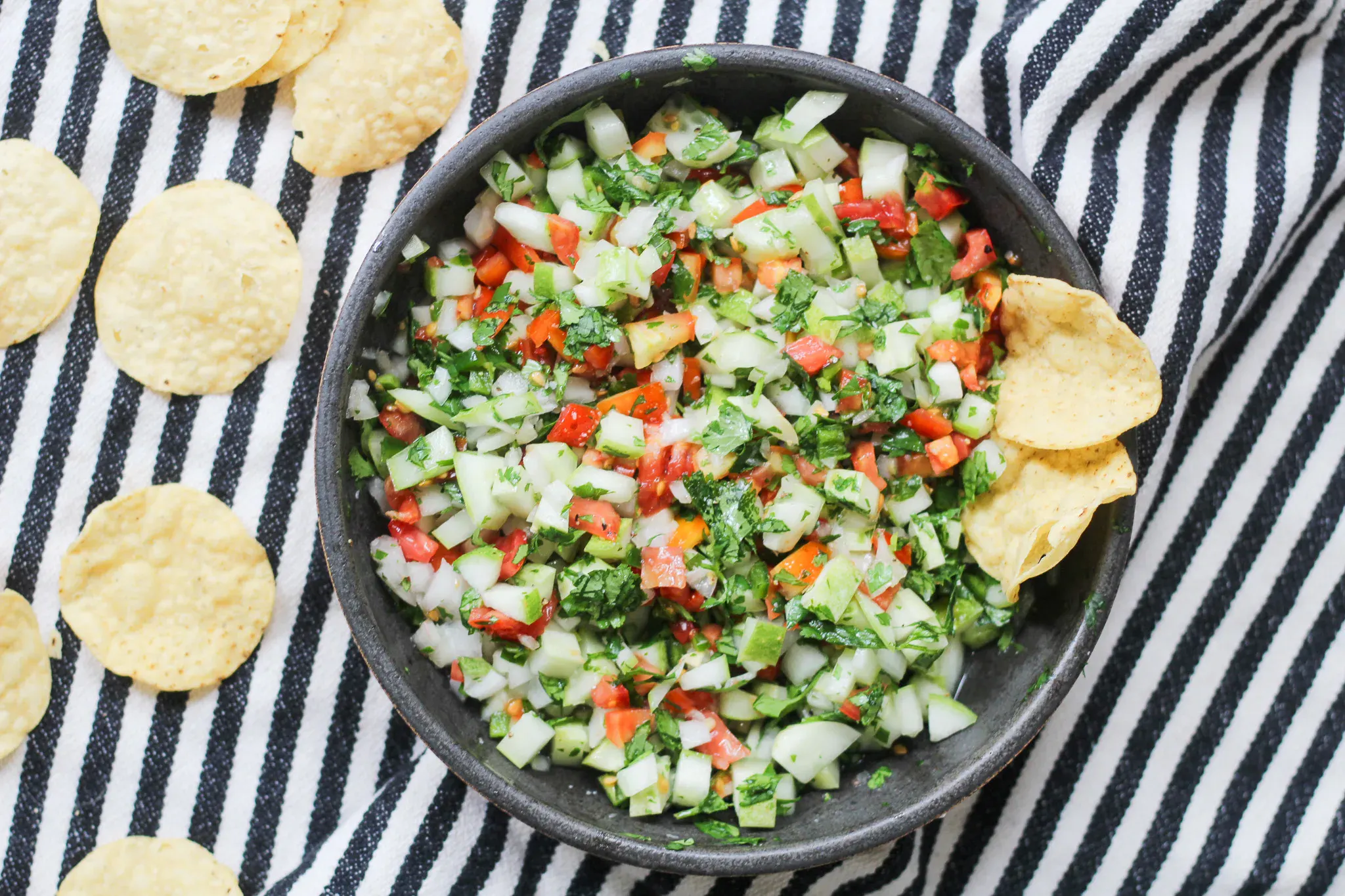 A bowl of fresh salsa with chopped tomatoes, cucumber, onion, and cilantro, surrounded by tortilla chips, sits on a black and white striped cloth.