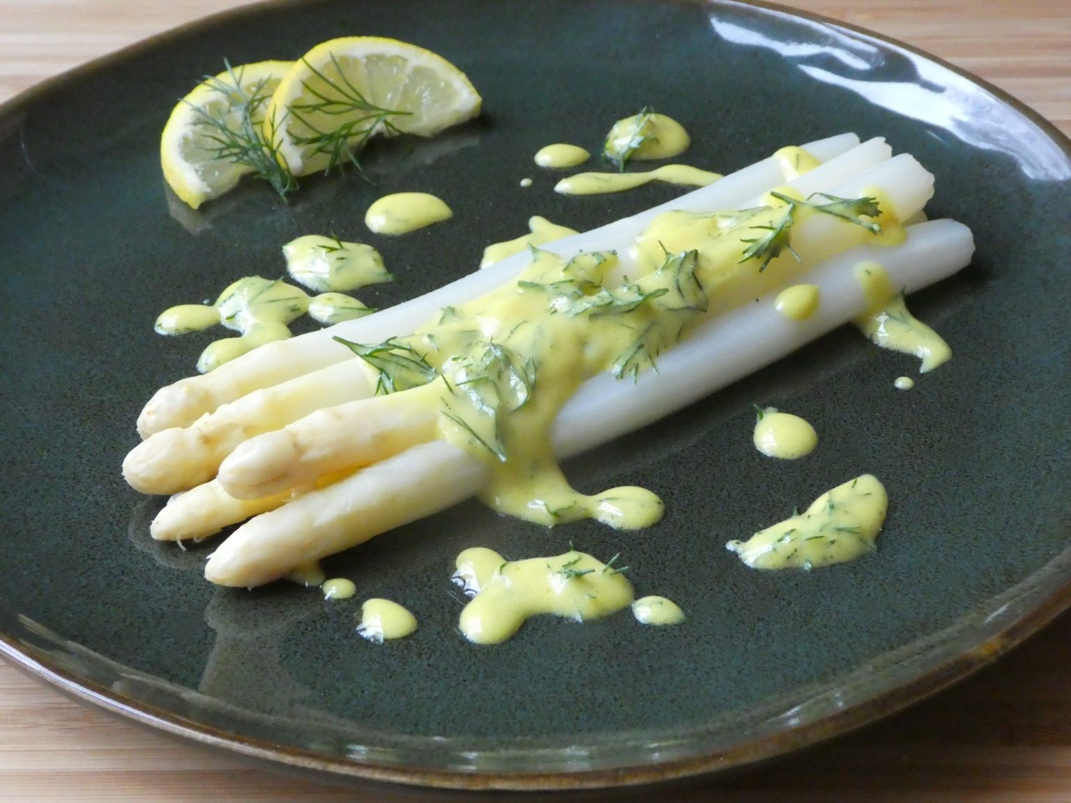 White asparagus spears topped with yellow sauce and dill, served on a dark plate with lemon wedges on the side.