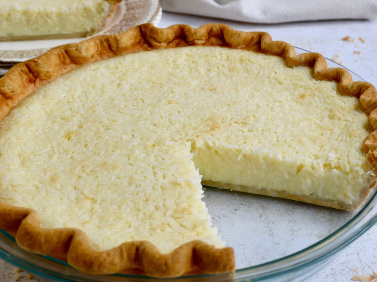 A baked coconut pie with a golden crust and creamy filling, displayed in a glass pie dish with one slice removed.
