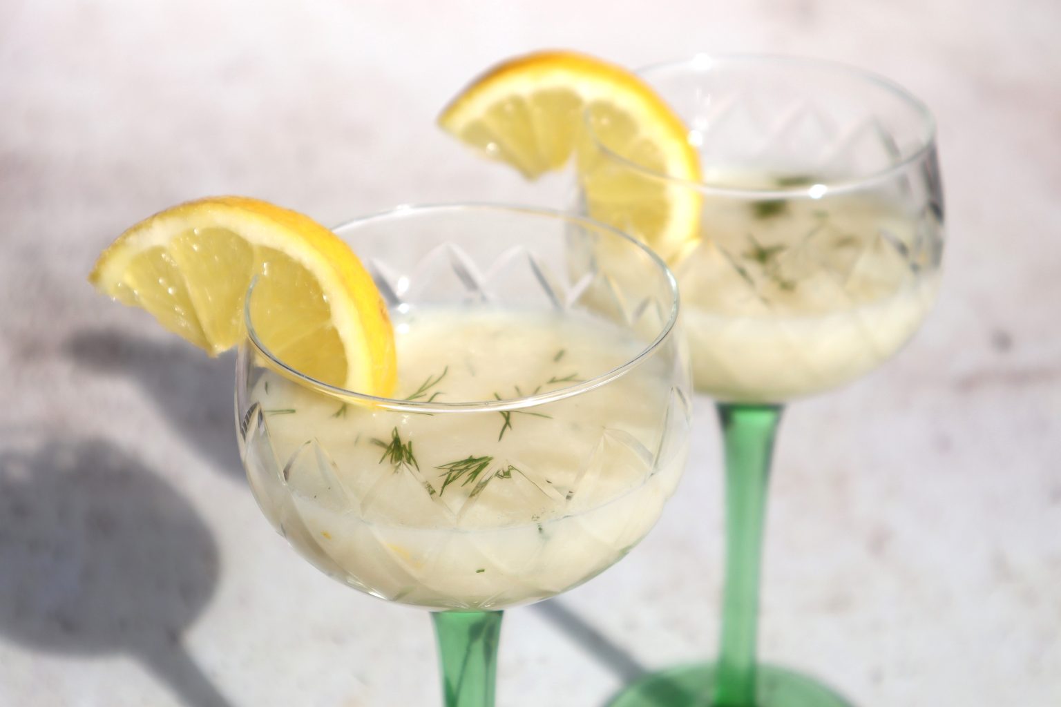 Two glasses with green stems contain a light-colored drink garnished with a lemon wedge and sprinkled herbs, placed on a white surface.