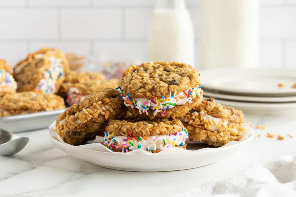 A plate of oatmeal cookie ice cream sandwiches with rainbow sprinkles on a white surface, with plates and bottles in the background.