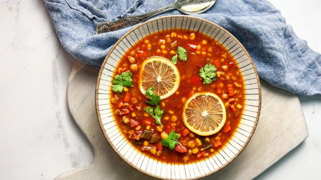 A bowl of tomato-based soup with corn, beans, lemon slices, and cilantro garnish, placed on a white cutting board next to a silver spoon and blue cloth.