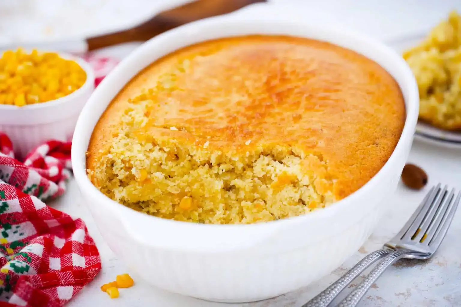 A baked cornbread casserole in a white dish with a portion scooped out, showing pieces of corn inside. A red checkered cloth and utensils are nearby.