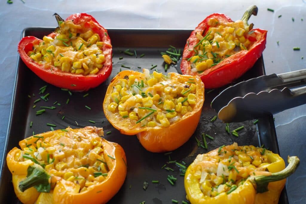 Bell pepper halves filled with a corn mixture are arranged on a black tray, garnished with chopped herbs. Metal tongs rest at the side of the tray.