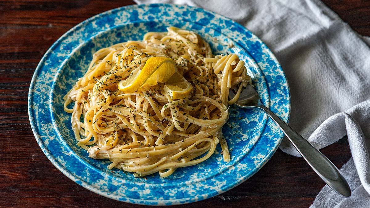 A plate of creamy pasta garnished with lemon wedges and herbs, served on a blue patterned dish with a fork, next to a white napkin on a wooden table.